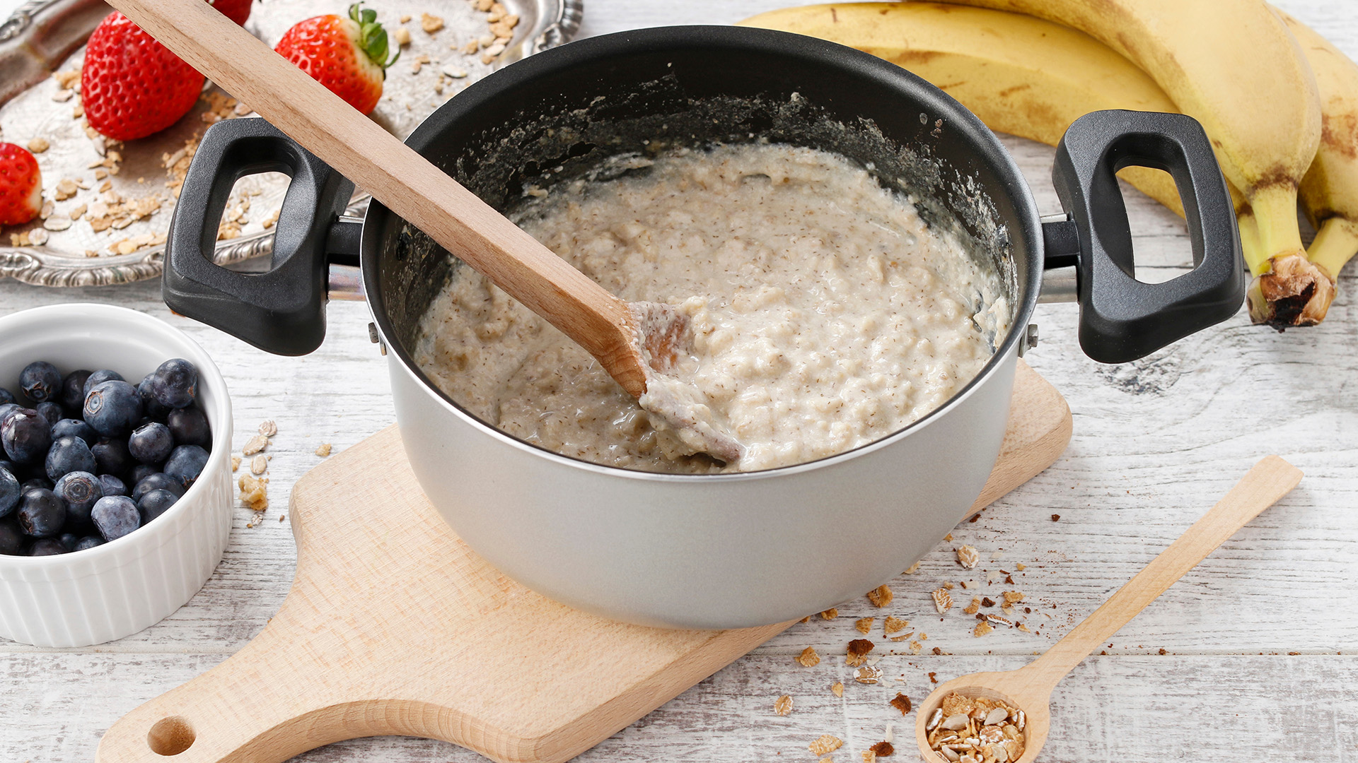 A photo of a pot of porridge with a wooden spoon surrounded by berries and nuts